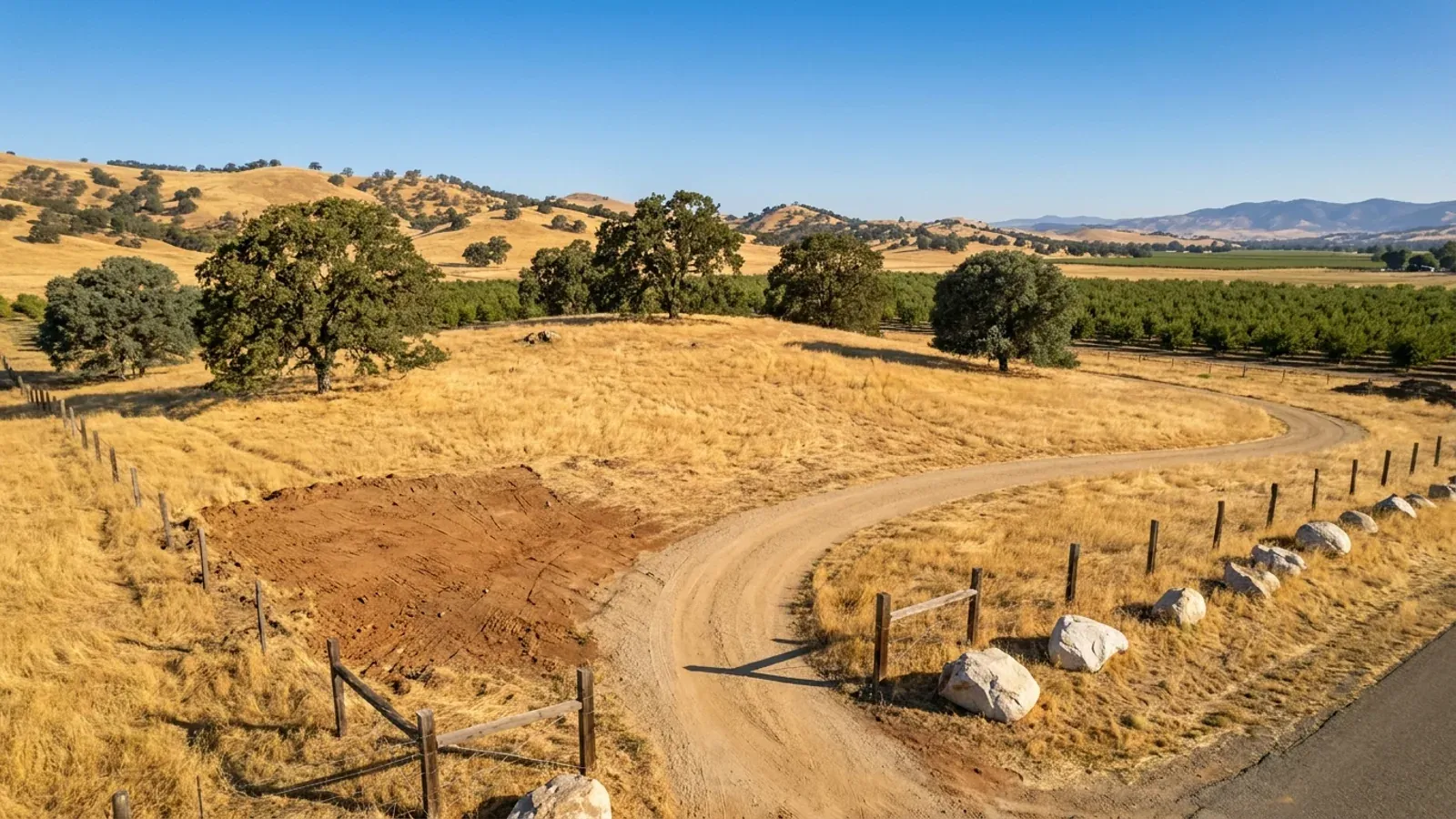 Aerial view of an undeveloped rural lot of golden dry grass with spreading oaks, granite boulders and a dirt access track, an orchard on the flat ground beyond.